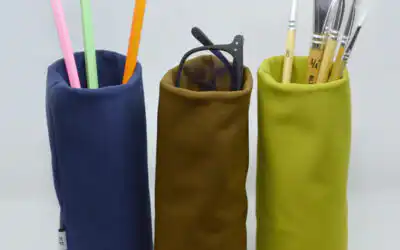 Three cylindrical fabric containers in blue, brown and green holding assorted pencils, paintbrushes and glasses.